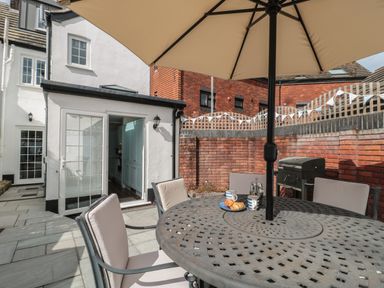 An outdoor patio with a round metal table and cushioned chairs under an umbrella outside a white house with an open door at Quay Cottage Sea View in Poole