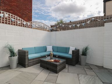 An outdoor patio area with a corner wicker sofa with blue cushions and a glass-top coffee table with a bottle of champagne two glasses and a cheese platter two potted plants and white brick walls with a wooden lattice fence and bunting at Quay Cottage Sea View in Poole
