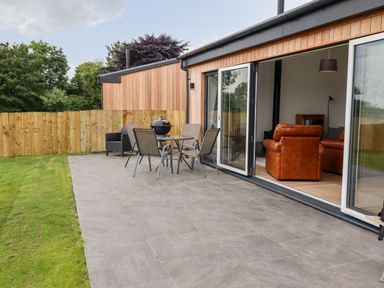 An outdoor patio with table and chairs next to a wooden building with sliding glass doors and brown leather chairs visible inside at 1 Orchard Cottages Dunstan near Craster
