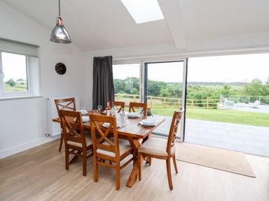 A dining room with a wooden table and six chairs near sliding glass doors opening to a patio and grassy yard at 1 Orchard Cottages Dunstan near Craster