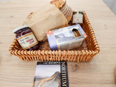 A wicker basket with bread jam marmalade and biscuits next to a book on a wooden table at 1 Orchard Cottages Dunstan near Craster