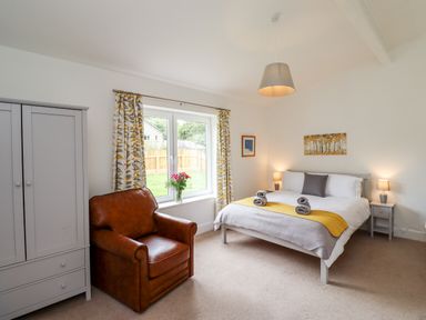 A bedroom with a bed nightstands with lamps a brown armchair a wardrobe a window with floral curtains and flowers on the windowsill at 1 Orchard Cottages in Dunstan near Craster