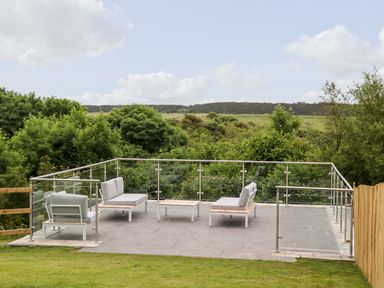 An outdoor patio with seating and tables surrounded by glass and metal railings and green trees at 1 Orchard Cottages in Dunstan near Craster