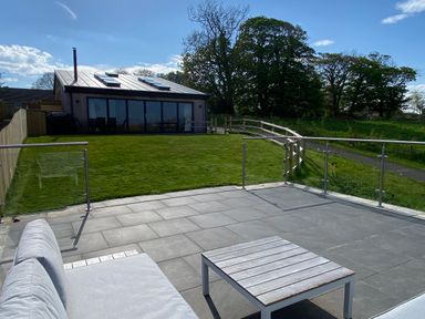 A patio with outdoor seating and a table overlooking a lawn and a modern house with skylights at 1 Orchard Cottages Dunstan near Craster