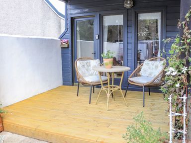 A wooden deck with two chairs and a round table with a potted plant in front of a cabin with blue walls and glass doors at Creed Cabin Stornoway