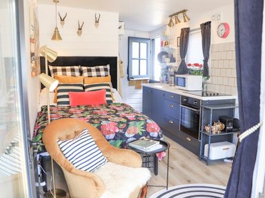 A small room with a bed with multiple pillows a floral quilt a kitchen counter with blue cabinets a microwave oven a chair with a striped cushion a floor lamp and wall decorations at Creed Cabin in Stornoway