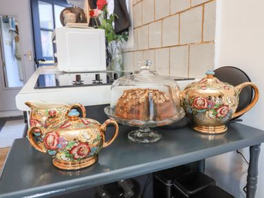 A kitchen counter with a stove a microwave vases with flowers and a table with a cake under a glass dome and floral teapot set at Creed Cabin in Stornoway