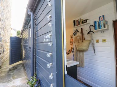 A blue exterior cabin wall with an open door showing a white interior wall with shelves holding books a hanging umbrella and a straw bag at Creed Cabin in Stornoway
