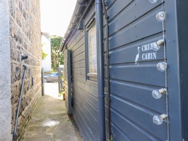 A narrow alley between a stone wall and a dark wooden cabin with garland lights and a sign saying Creed Cabin in Stornoway