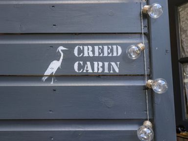A dark wooden exterior wall with a white bird stencil and text Creed Cabin and string lights attached at Creed Cabin in Stornoway