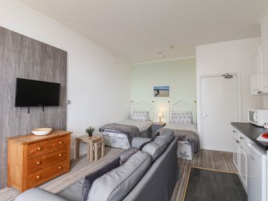 A room with two single beds a sofa a wooden chest of drawers a wall-mounted TV and a kitchenette at 4 Beach View Beaconsfield House in Bridlington