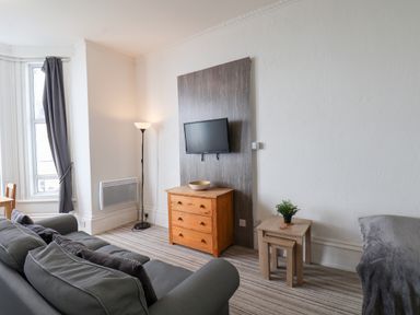 A living room with a gray sofa a wooden chest of drawers a small table with a plant a floor lamp and a wall-mounted TV at 4 Beach View Beaconsfield House Bridlington