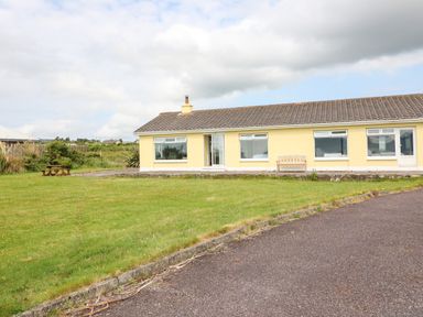 A single-story yellow house with a tiled roof and multiple windows on a grassy lawn with a paved driveway at Meeligolleen in Coill Tra