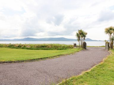 A paved driveway leading to a gate with palm trees beside a grassy area and a body of water with hills in the background at Meeligolleen in Coill Tra
