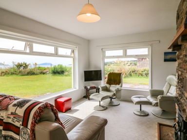 A living room with large windows showing a garden and hills with white leather chairs and a stone fireplace at Meeligolleen in Coill Tra