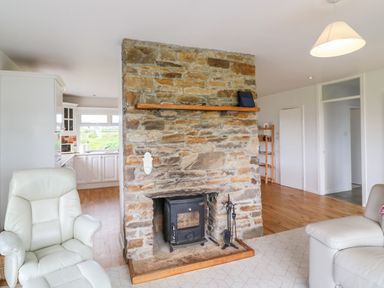 A room with a stone fireplace and wood stove between a kitchen and a living area with white chairs at Meeligolleen in Coill Tra