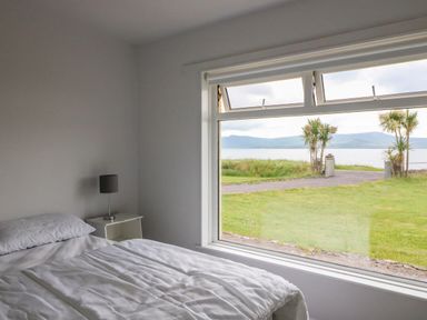 A bedroom with a bed side table and lamp next to a large window overlooking a grassy area with palm trees and water at Meeligolleen in Coill Tra