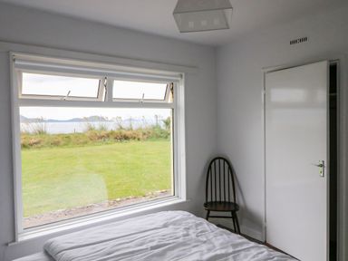 A bedroom with a bed near a large window overlooking grass and water with a wooden chair and a closed door at Meeligolleen in Coill Tra