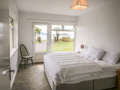 A bedroom with a bed pillows bedside table and chair next to a door with a window showing grass and water outside at Meeligolleen in Coill Tra