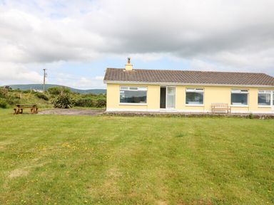 A yellow single-story house with large windows and a bench on a grassy lawn at Meeligolleen in Coill Tra