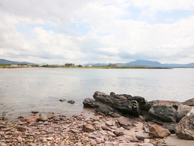A rocky shoreline with water and distant buildings with mountains in the background at Meeligolleen in Coill Tra