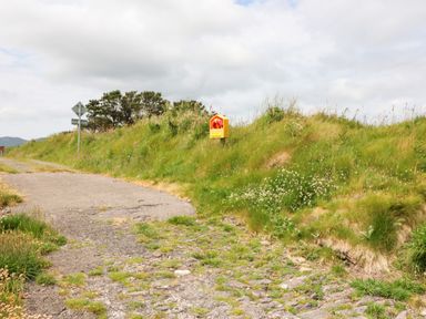 A rural roadside with a paved and rocky path and grassy embankment with a yellow emergency box and traffic signs at Meeligolleen in Coill Tra