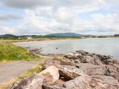 A rocky shoreline beside a calm bay with grass and houses in the distance at Meeligolleen in Coill Tra