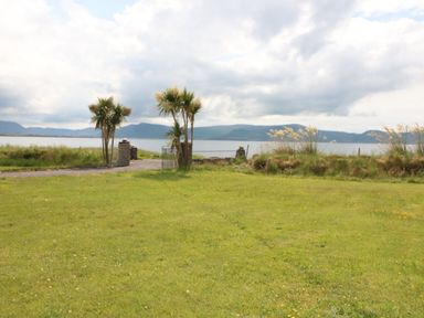 A grassy area with palm trees and a metal gate overlooking water and mountains at Meeligolleen in Coill Tra