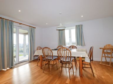 A dining room with wooden chairs around a table covered with a tablecloth a high chair to the side and windows with striped curtains at Freefolk in Polzeath