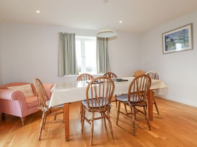 A dining room with a wooden table covered with a tablecloth surrounded by six wooden chairs a pink armchair near the window with curtains and a framed picture on the wall at Freefolk in Polzeath
