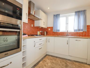 A kitchen with white cabinets orange tile backsplash and a window with blue check curtains at Freefolk in Polzeath