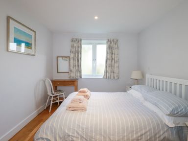A bedroom with a bed with striped bedding towels on the bed a wooden chair and desk near a window with floral curtains and a lamp on a side table at Freefolk in Polzeath