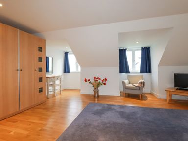 A room with wooden floor a wardrobe a chair a desk with a mirror a television on a wooden stand and windows with blue curtains at Freefolk in Polzeath