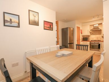 A dining area with a wooden table and six chairs near a kitchen with appliances and framed pictures on the wall at The Mill Stones in Stalham