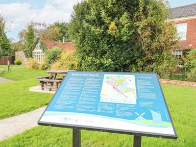 A trail information board with a map and text next to wooden picnic benches and houses with trees and grass at The Mill Stones in Stalham