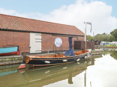 A wooden boat docked beside a brick building with a red tiled roof by the water at The Mill Stones in Stalham