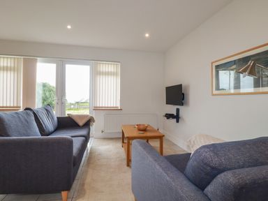 A living room with two blue sofas a wooden coffee table a wall-mounted TV and glass doors with blinds at Freefolk Cottage Polzeath