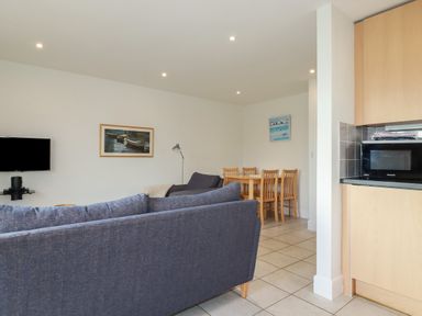 A living room with blue sofas a wall-mounted television a wooden dining table with chairs and a microwave on a kitchen counter at Freefolk Cottage Polzeath