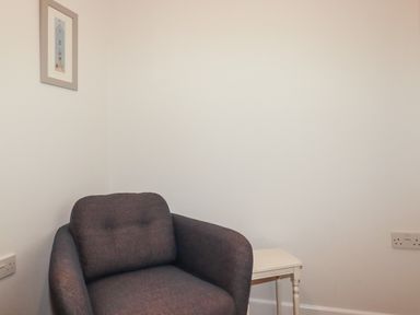A corner of a room with a gray armchair a small white side table and a framed picture on the wall at Freefolk Cottage Polzeath