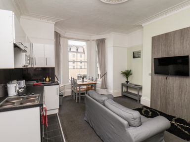 An open plan kitchen and living room with a gray sofa a wall mounted television a dining table with chairs and large windows at Promenade in Bridlington
