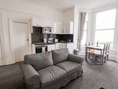 A living area with a gray sofa, a kitchen with white cabinets and appliances, and a dining table with four chairs by large windows at Promenade Bridlington
