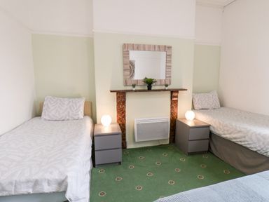 A bedroom with two single beds, two nightstands with lamps, a mirror above a mantle with plants, and green patterned carpet at Promenade in Bridlington