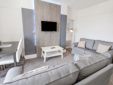 A living room with grey sofas a white coffee table a wall-mounted TV and a dining table with white chairs at Ocean Wave in Bridlington