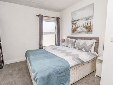 A bedroom with a bed with chevron patterned bedding a blue throw a window with curtains and a beach painting above the bed at Ocean Wave Bridlington