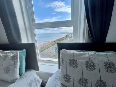 A window view of the sea and road between two beds with patterned pillows and curtains at Ocean Wave in Bridlington