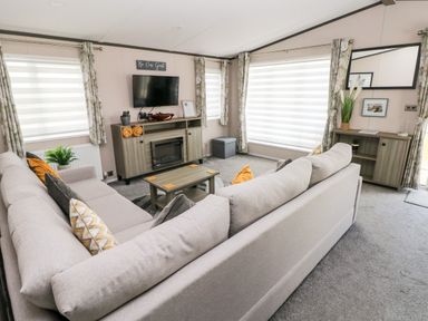 A living room with two gray sofas a wooden TV stand with a fireplace a wall-mounted TV and large windows with curtains at Beechwood Lodge in Hasguard Cross near Broad Haven