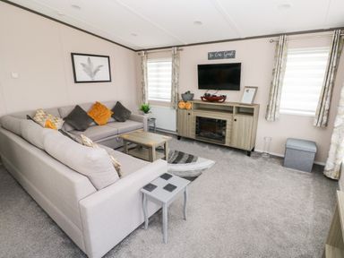 A living room with a sectional sofa, coffee table, television on a stand, and two windows with curtains at Beechwood Lodge in Hasguard Cross near Broad Haven