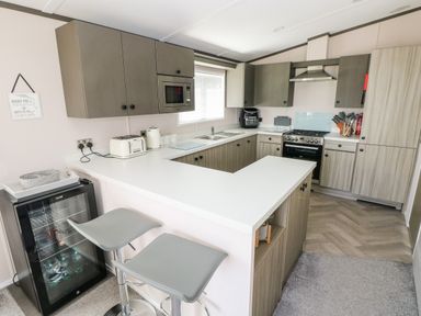 A kitchen with grey cabinets white countertops bar stools and a small fridge at Beechwood Lodge in Hasguard Cross near Broad Haven