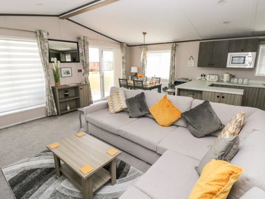 A living room with a light gray sectional sofa with pillows a wooden coffee table a patterned rug and a dining table with chairs near kitchen cabinets at Beechwood Lodge Hasguard Cross near Broad Haven