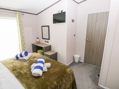 A bedroom with a bed covered by a brown blanket and towels on top a desk with a mirror a TV mounted on the wall a door and a window with patterned curtains at Beechwood Lodge Hasguard Cross near Broad Haven